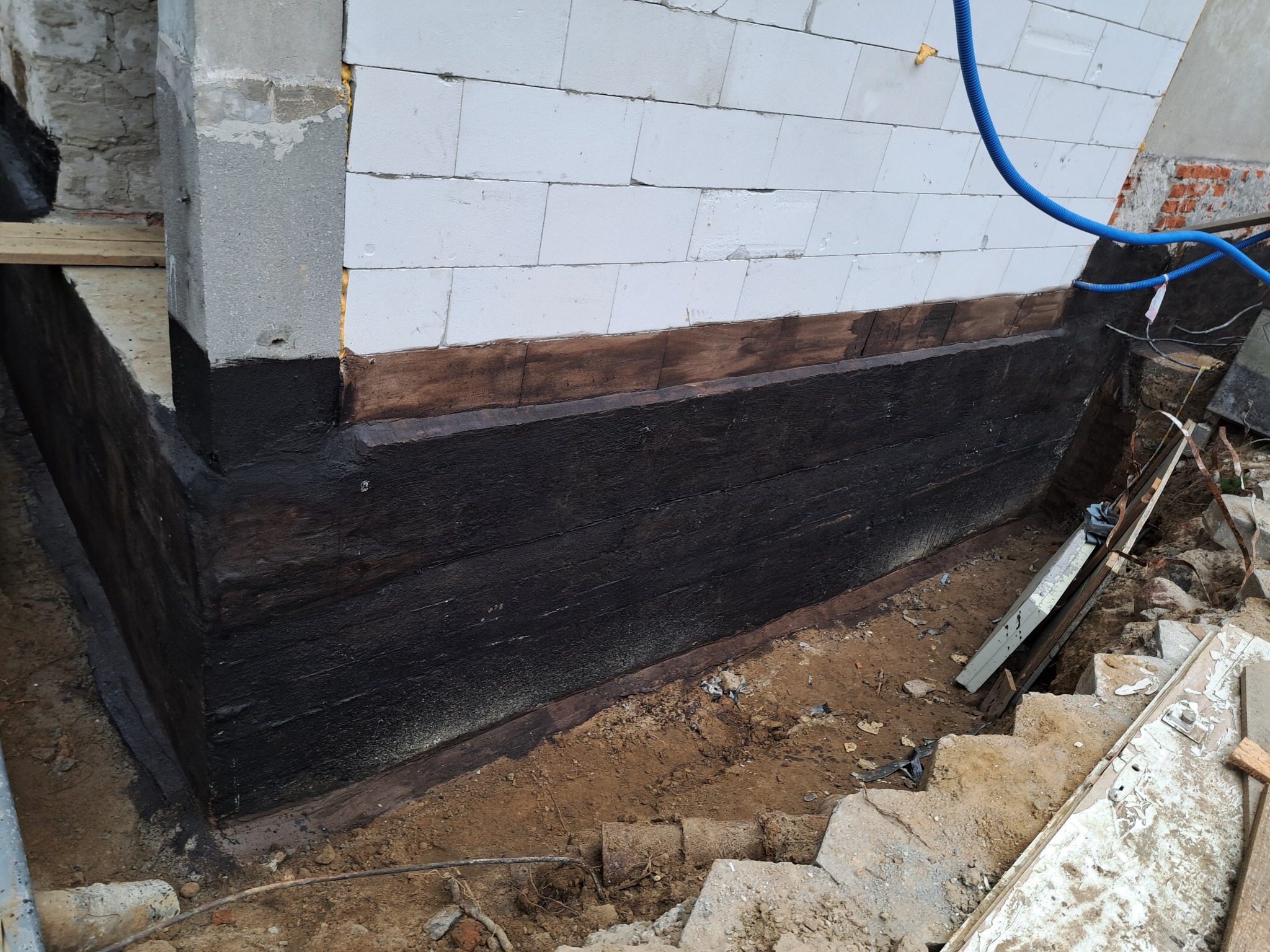 Zabezpieczona czarną powłoką hydroizolacyjną zewnętrzna ściana fundamentowa z bloczków betonowych, częściowo zakopana w ziemi, widoczne elementy deskowania i niebieski przewód ochronny.