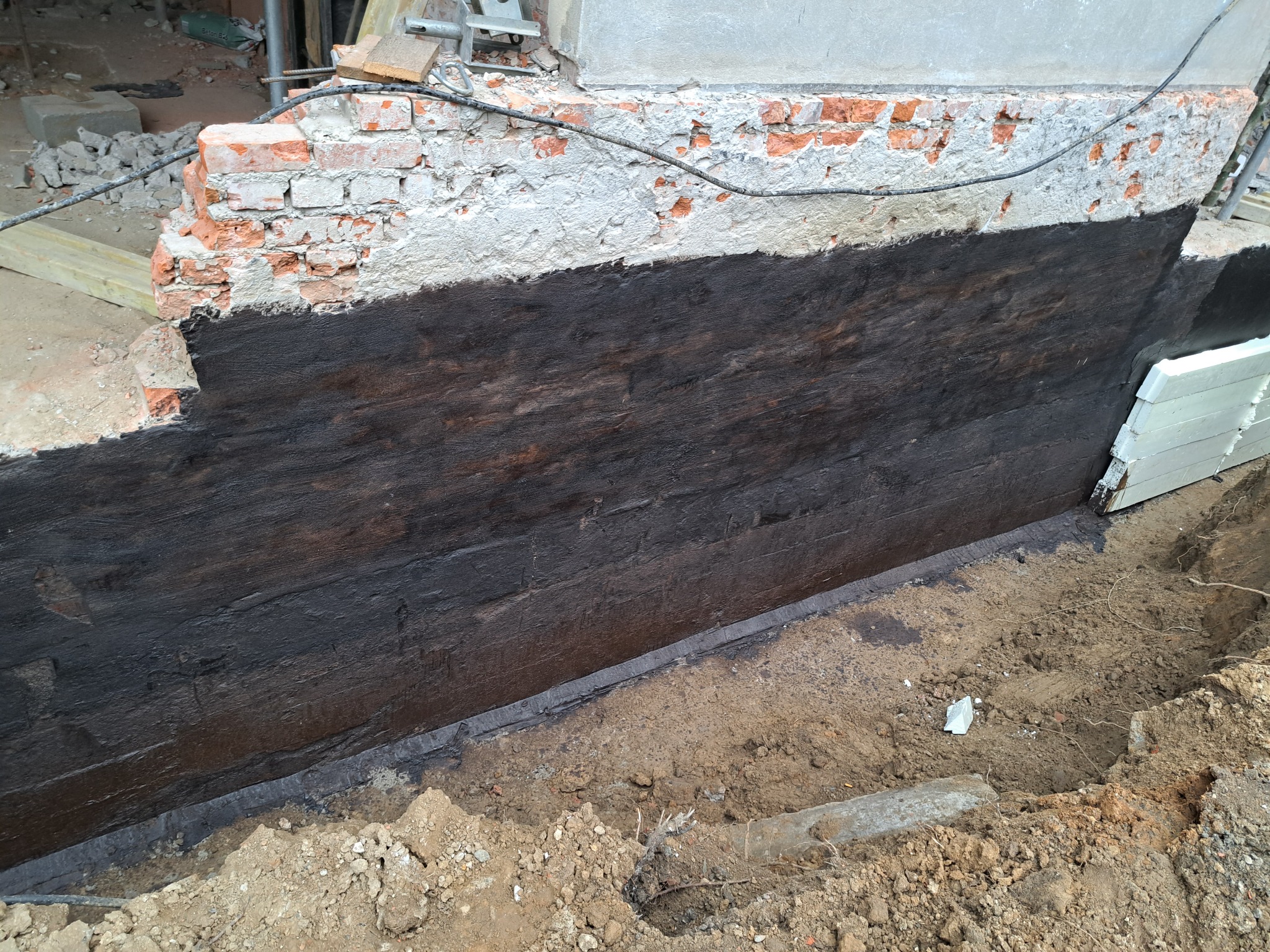 Świeżo zabezpieczona, czarną powłoką hydroizolacyjną, ściana fundamentowa z fragmentami cegieł i betonu, widoczna w wykopie ziemnym.