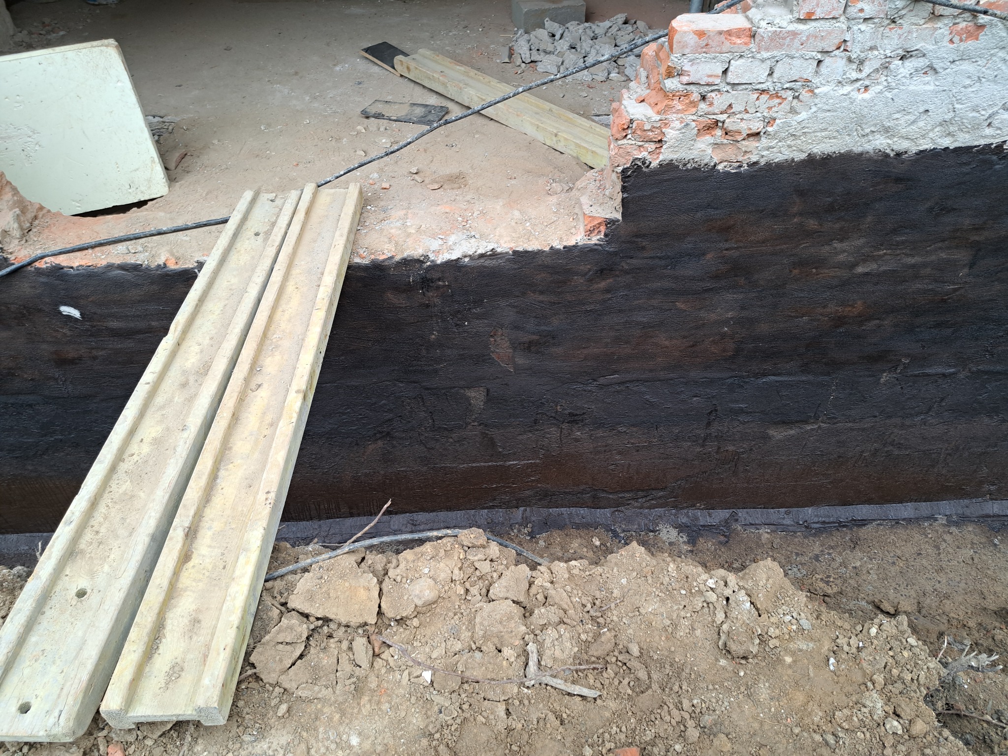 Świeżo zabezpieczona czarną powłoką ściana fundamentowa, z drewnianymi deskami opartymi o nią i fragmentami cegieł na górze, ukazująca etap prac hydroizolacyjnych.