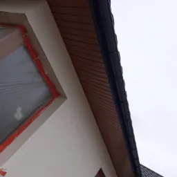 Fragment elewacji domu z oknem zabezpieczonym folią, świeżo otynkowaną ścianą i drewnianą podbitką dachową z zamontowaną rynną