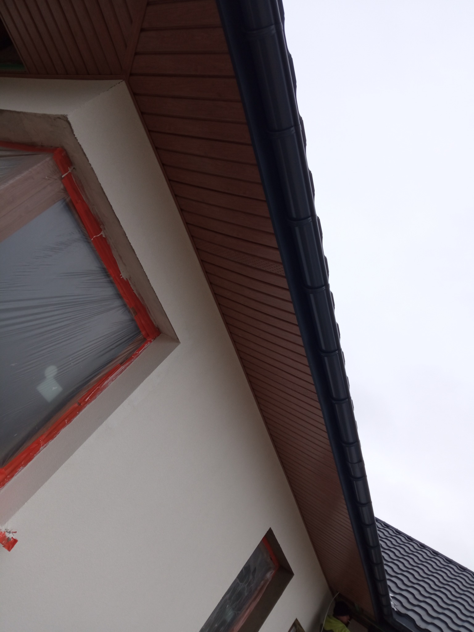 Fragment elewacji domu z oknem zabezpieczonym folią, świeżo otynkowaną ścianą i drewnianą podbitką dachową z zamontowaną rynną