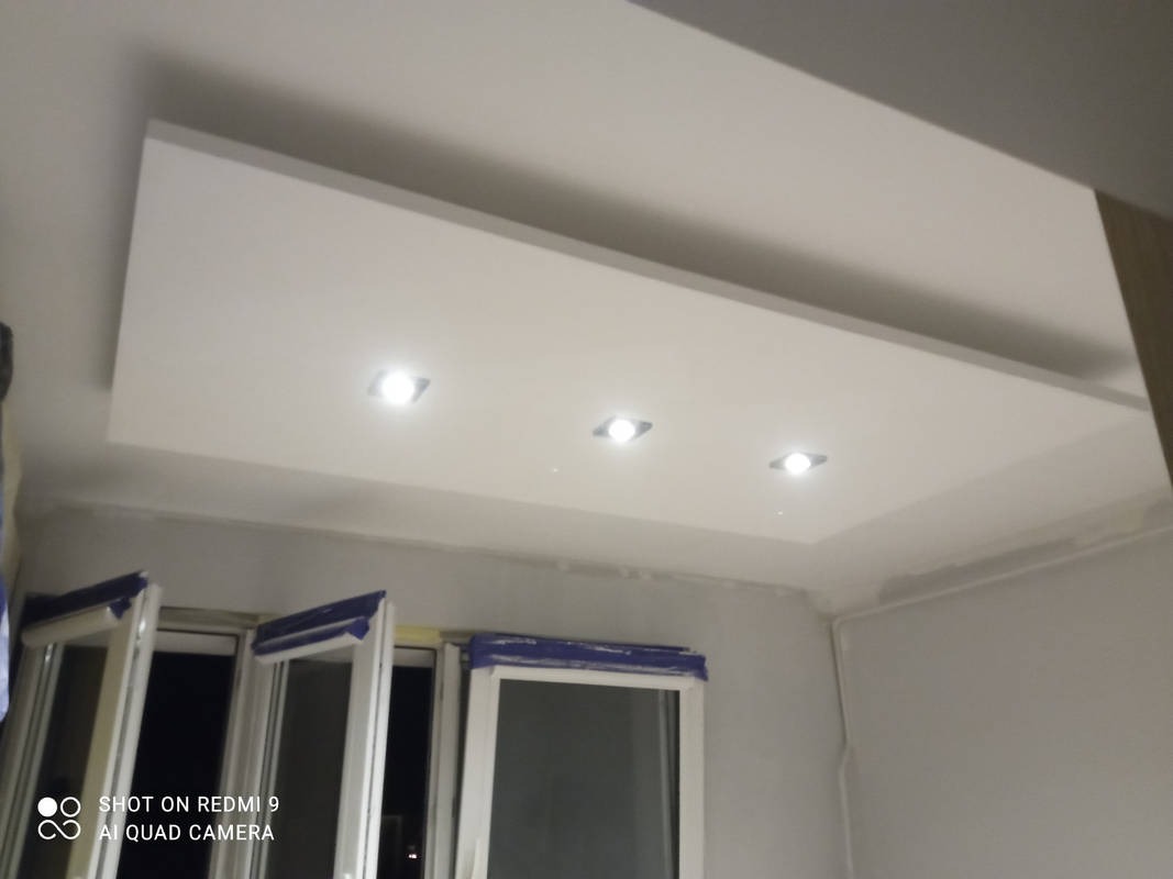 Biały sufit podwieszany z trzema wbudowanymi punktami świetlnymi, widoczne okna zabezpieczone folią malarską, ściany w trakcie wykańczania.