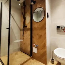 Nowoczesna łazienka z kabiną prysznicową typu walk-in z czarnymi ramami, drewnopodobne płytki ułożone w jodełkę na ścianie i podłodze, okrągłe lustro na pasku i czarne lampy ścienne.