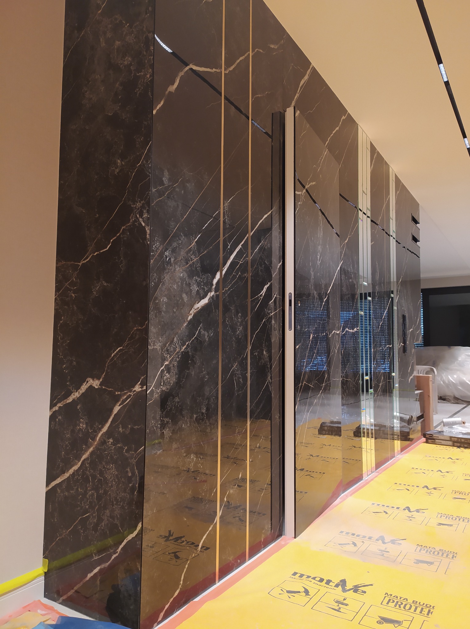Ściana wykończona panelami imitującymi czarny marmur z białymi żyłami, oddzielonymi cienkimi złotymi paskami, z widoczną ochronną folią na podłodze.