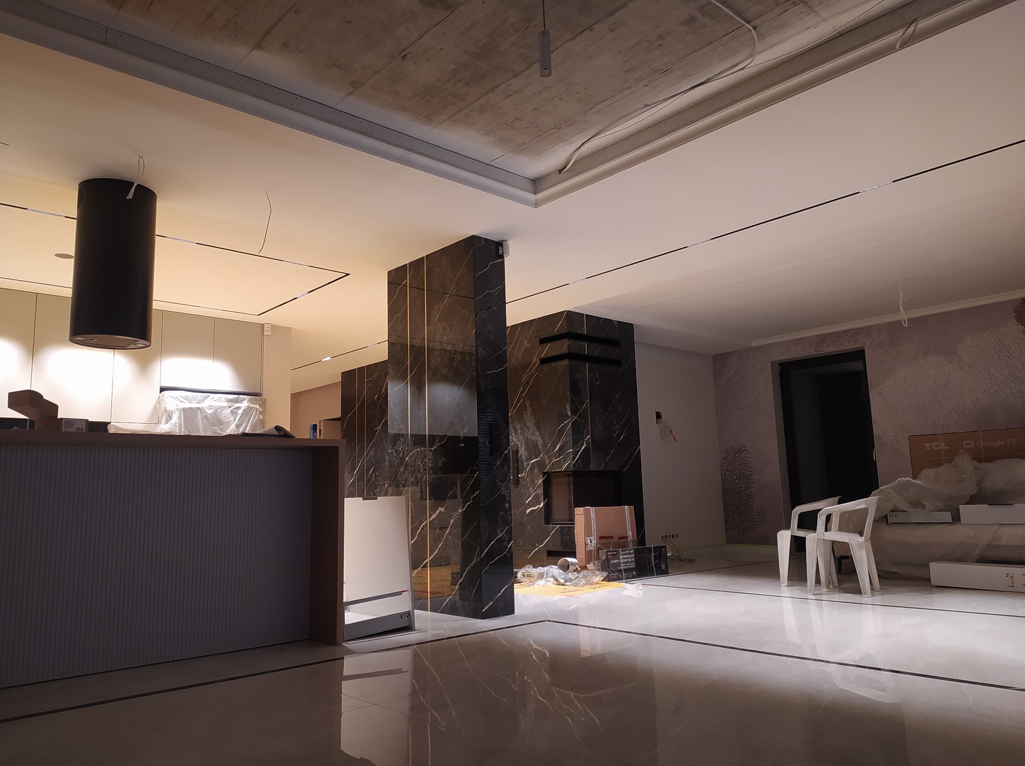 Wnętrze w trakcie wykańczania: podwieszany sufit z oświetleniem liniowym, ściana z czarnego marmuru z kominkiem, białe krzesła i kartony na podłodze.
