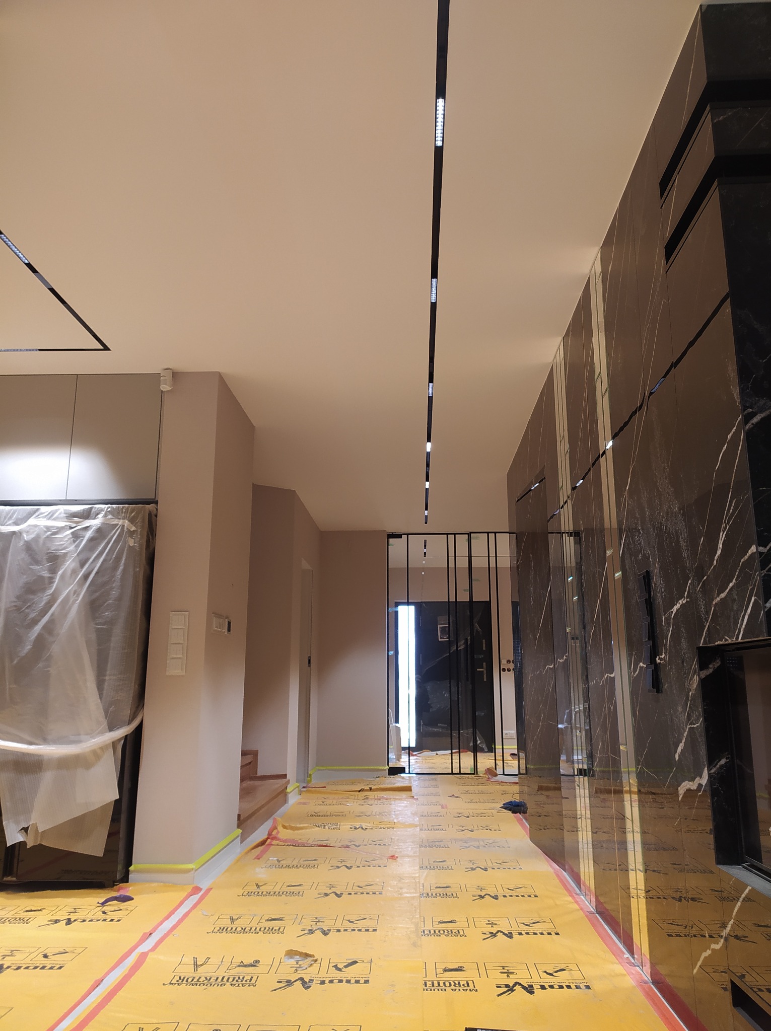 Wnętrze w trakcie remontu: zabezpieczona podłoga folią ochronną, ściana wykończona czarnym marmurem z białymi żyłami, widoczne listwy oświetleniowe w suficie.
