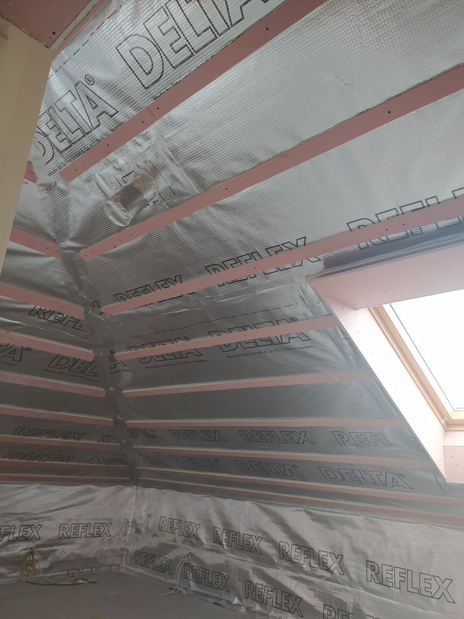 Montaż paroizolacji Delta Reflex na poddaszu, widoczne srebrzysta folia z nadrukiem, drewniane łaty i fragment okna dachowego.