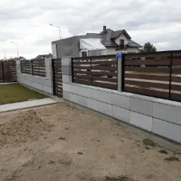 Nowoczesne ogrodzenie z betonowych bloczków i paneli imitujących drewno, widoczny stos piasku na jeszcze niezagospodarowanym terenie, w tle dom jednorodzinny i samochód dostawczy.