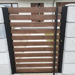 Nowoczesna brama wjazdowa z poziomymi panelami drewnopodobnymi, osadzona w czarnych metalowych ramach i murowanych słupkach z betonu architektonicznego, widok na ścieżkę i elewację domu w tle.