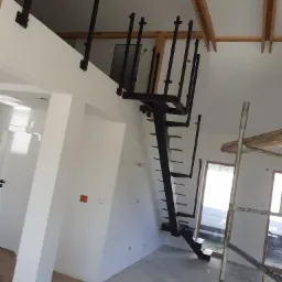 Surowe, czarne schody metalowe bez balustrady, zamontowane w nowoczesnym wnętrzu w trakcie wykańczania, widoczne drewniane belki stropowe i rusztowanie.