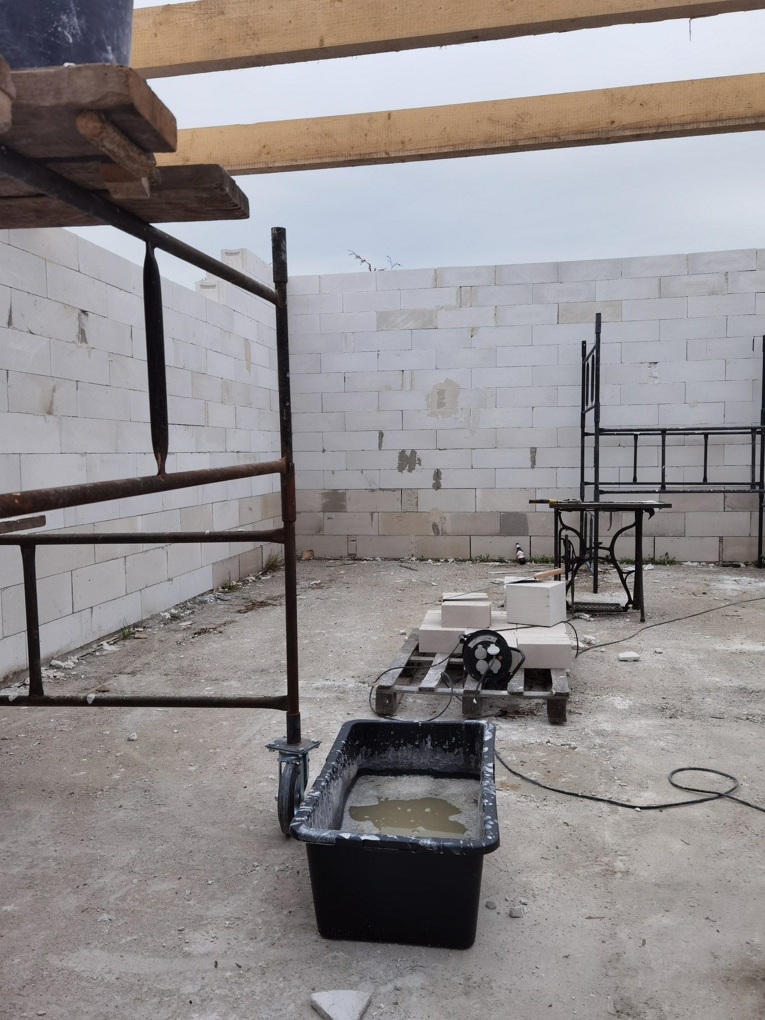 Wznoszenie ściany z białych bloczków betonowych na placu budowy, widoczne rusztowanie, paleta z bloczkami, wiadro z zaprawą i prowizoryczny stół roboczy.