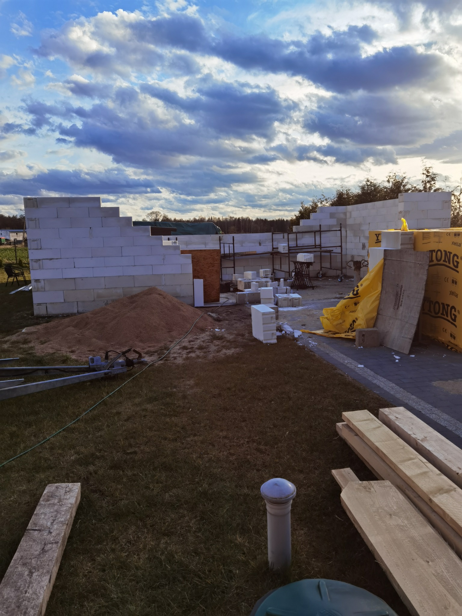 Budowa domu jednorodzinnego w trakcie: wznoszone ściany z bloczków, widoczny piasek, materiały budowlane i rusztowanie na tle zachmurzonego nieba.