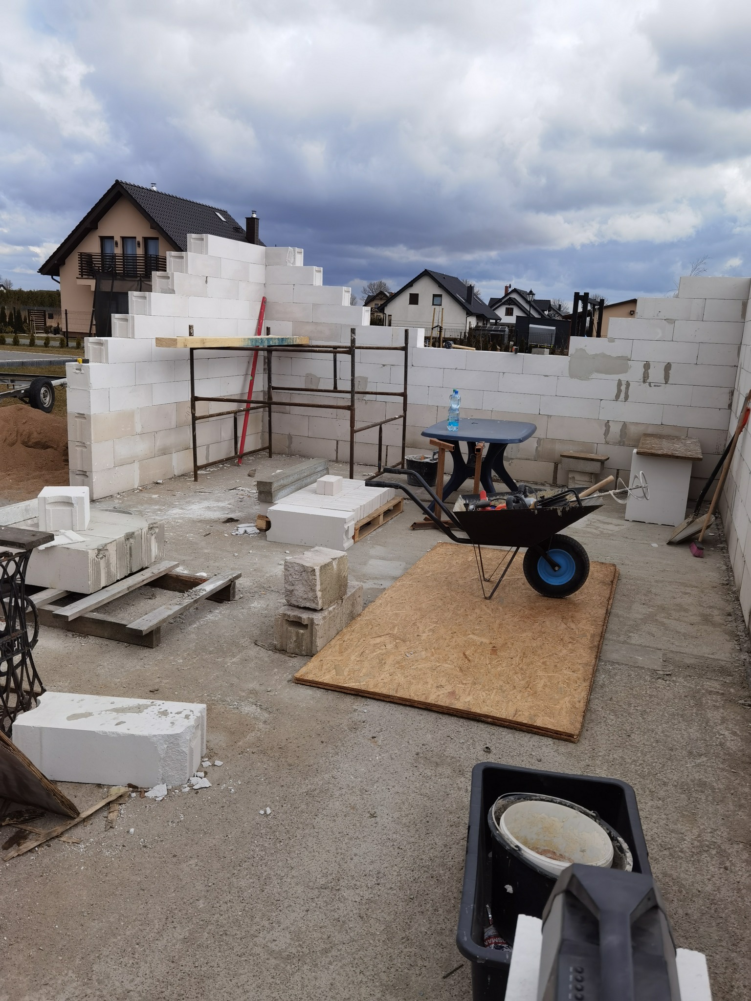 Wznoszenie ścian budynku z białych bloczków, widoczne rusztowanie, taczka z narzędziami, butelka wody na stole i dom w tle pod pochmurnym niebem.