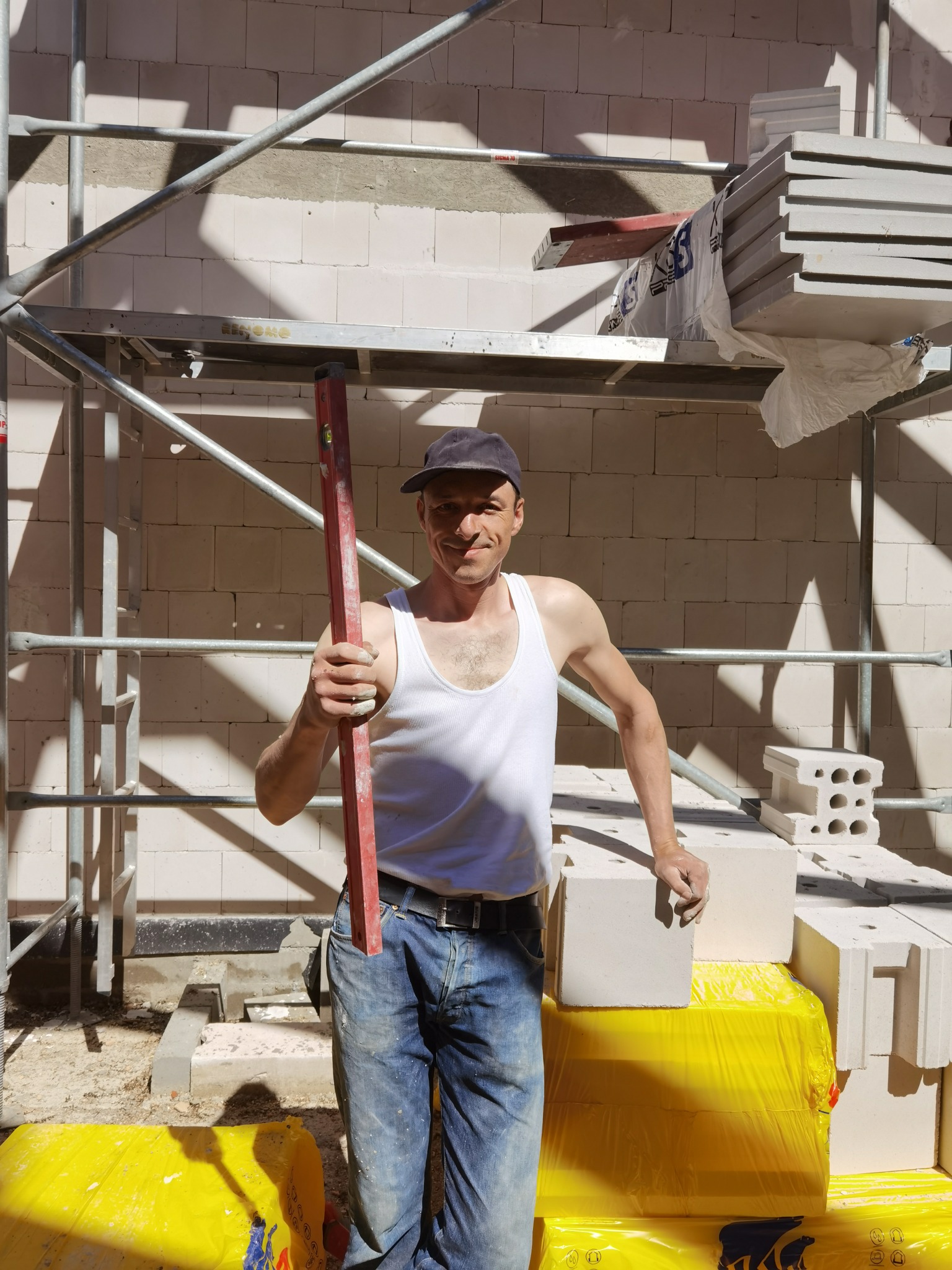 Murarz w podkoszulku i czapce, stojący na rusztowaniu z poziomicą w ręku, obok bloczków z betonu komórkowego na żółtej folii, w tle ściana z białych bloczków.