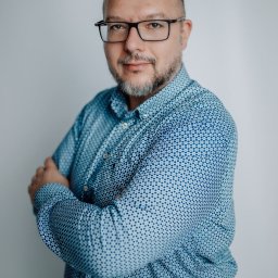 Libera Mentis - Marcin Żekoński - Portret mężczyzny w okularach i niebieskiej koszuli w kropki, z rękami skrzyżowanymi na piersiach, na jasnym tle.