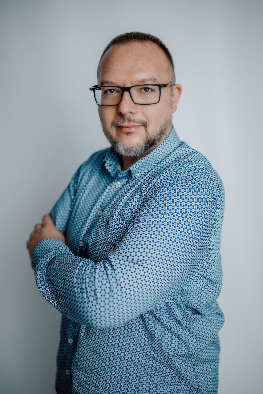 Portret mężczyzny w okularach i niebieskiej koszuli w kropki, z rękami skrzyżowanymi na piersiach, na jasnym tle.