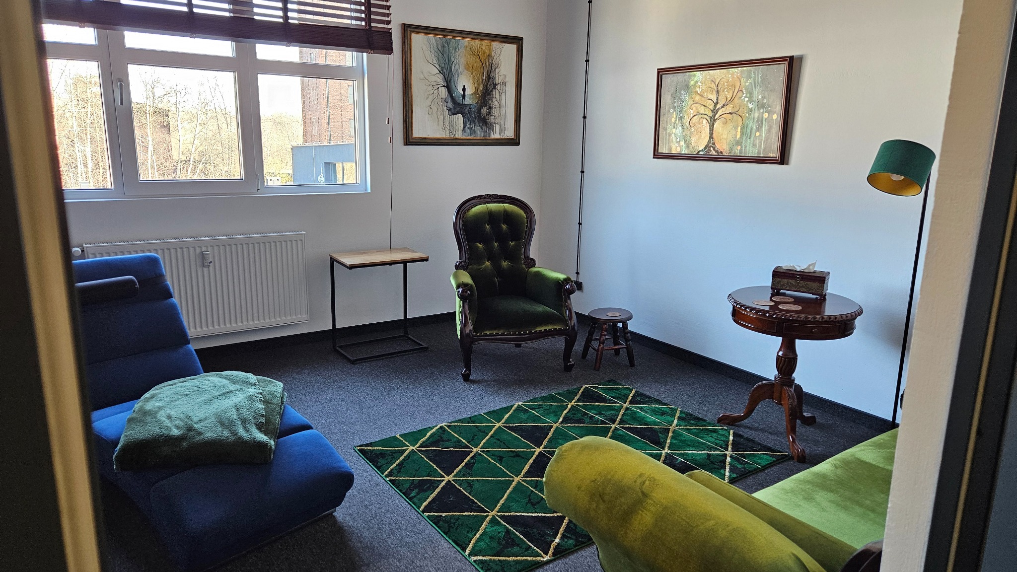 Gabinet terapeutyczny w Chorzowie z zielonymi akcentami: fotel typu tron, dywan w geometryczne wzory, drewniany stolik z chusteczkami, obrazy na ścianach i niebieski leżak relaksacyjny.
