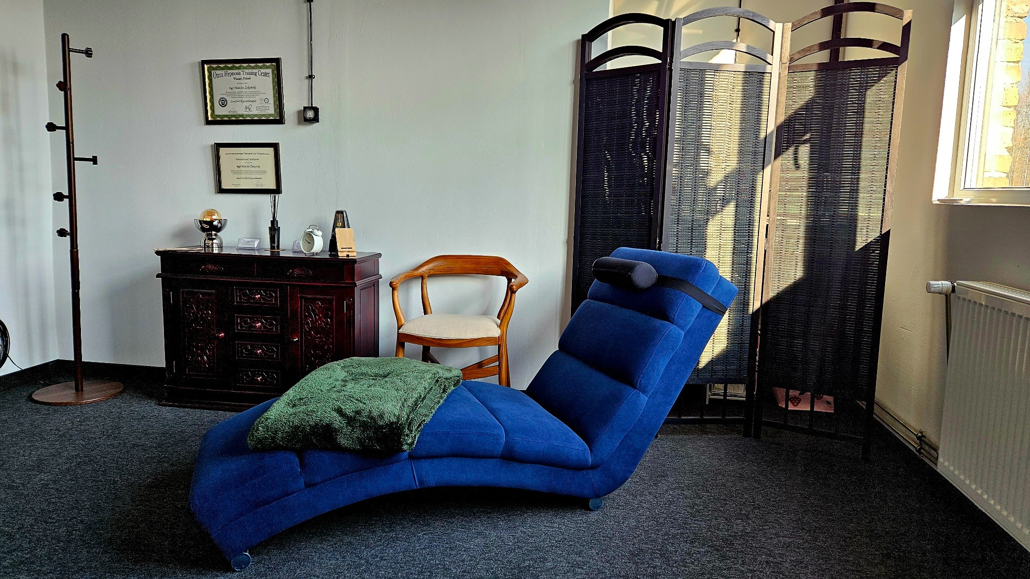 Pokój z niebieskim szezlongiem z zagłówkiem, przykrytym zielonym kocem, drewnianym krzesłem, parawanem, komodą z dekoracjami i certyfikatami na ścianie.