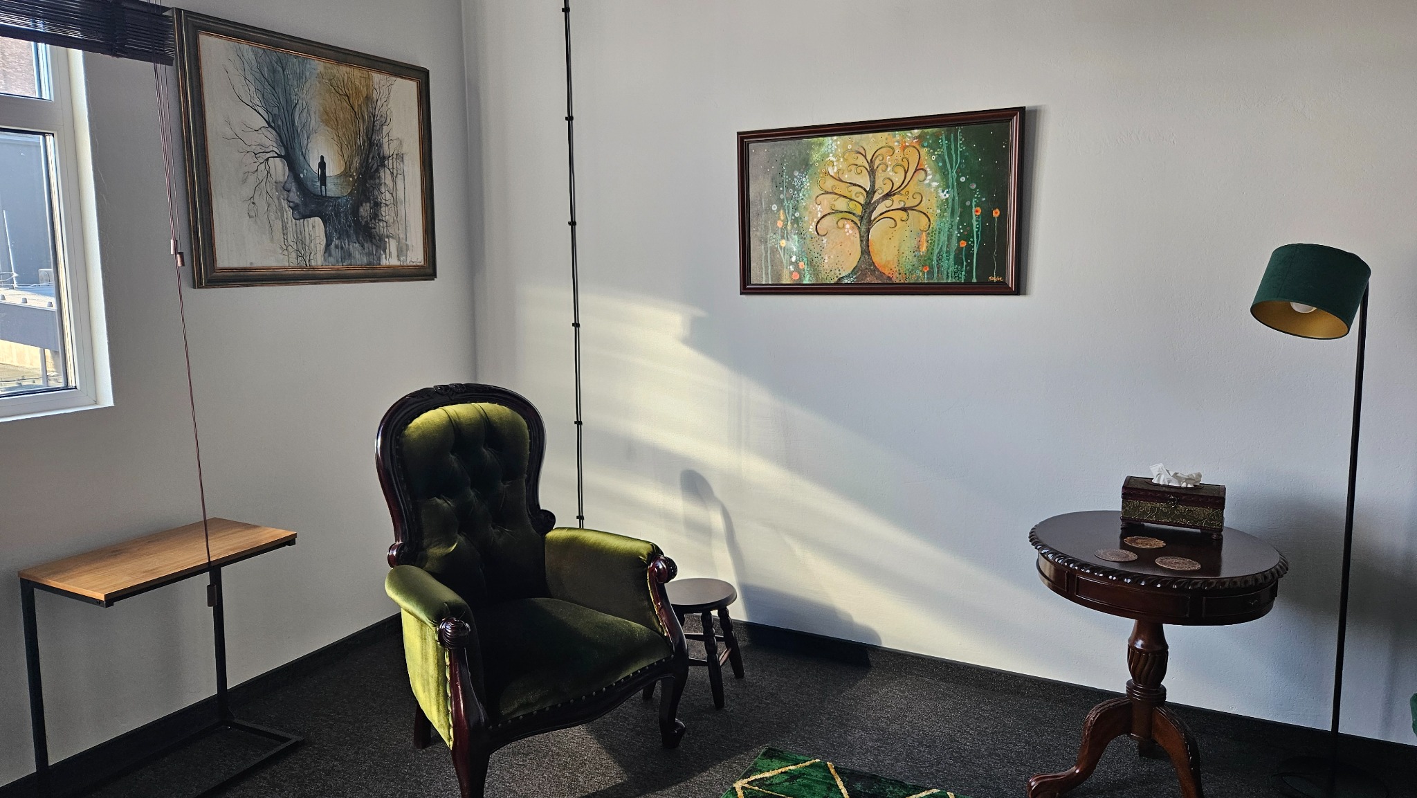 Pokój z zielonym, aksamitnym fotelem, stolikiem z lampą i dwoma obrazami z motywami drzew na białej ścianie, oświetlony promieniami słonecznymi.