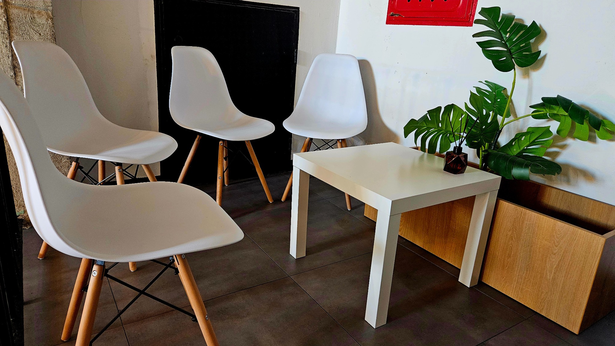 Aranżacja poczekalni: cztery białe krzesła Eames z drewnianymi nogami, kwadratowy biały stolik z brązowym dyfuzorem zapachowym i zielona monstera w drewnianej osłonce na tle białej ściany...