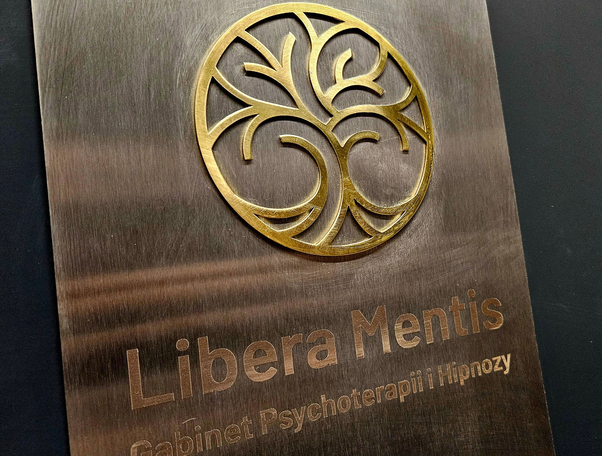 Metalowa tabliczka z logo drzewa życia i napisem 'Libera Mentis Gabinet Psychoterapii i Hipnozy', złote elementy na szczotkowanym tle.