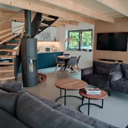 Przestronny salon z drewnianymi panelami ściennymi i sufitem, spiralnymi schodami z drewnianymi stopniami, nowoczesnym piecem wolnostojącym, szarą sofą i fotelami oraz otwartą kuchnią z niebieskimi...