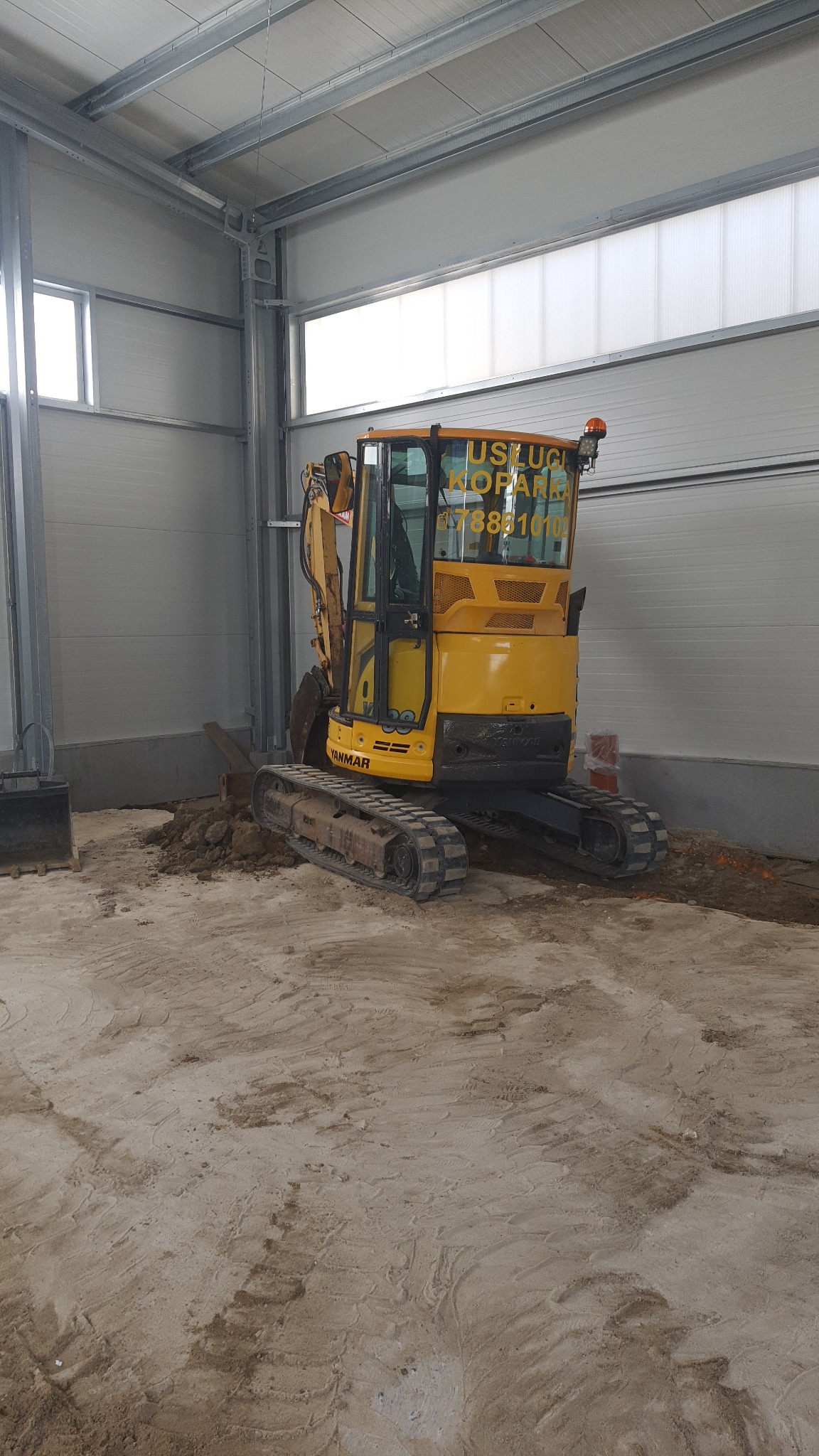 Żółta mini koparka Yanmar z numerem telefonu na szybie kabiny stoi wewnątrz hali z betonową posadzką, obok wykopany rów.