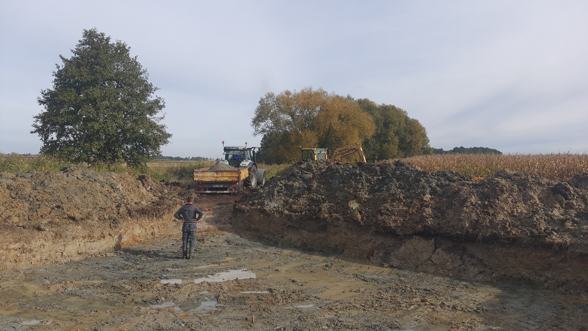 Mężczyzna w ubraniu roboczym stoi przy wykopie w grząskim terenie, w tle traktor z przyczepą, koparka i pole kukurydzy.
