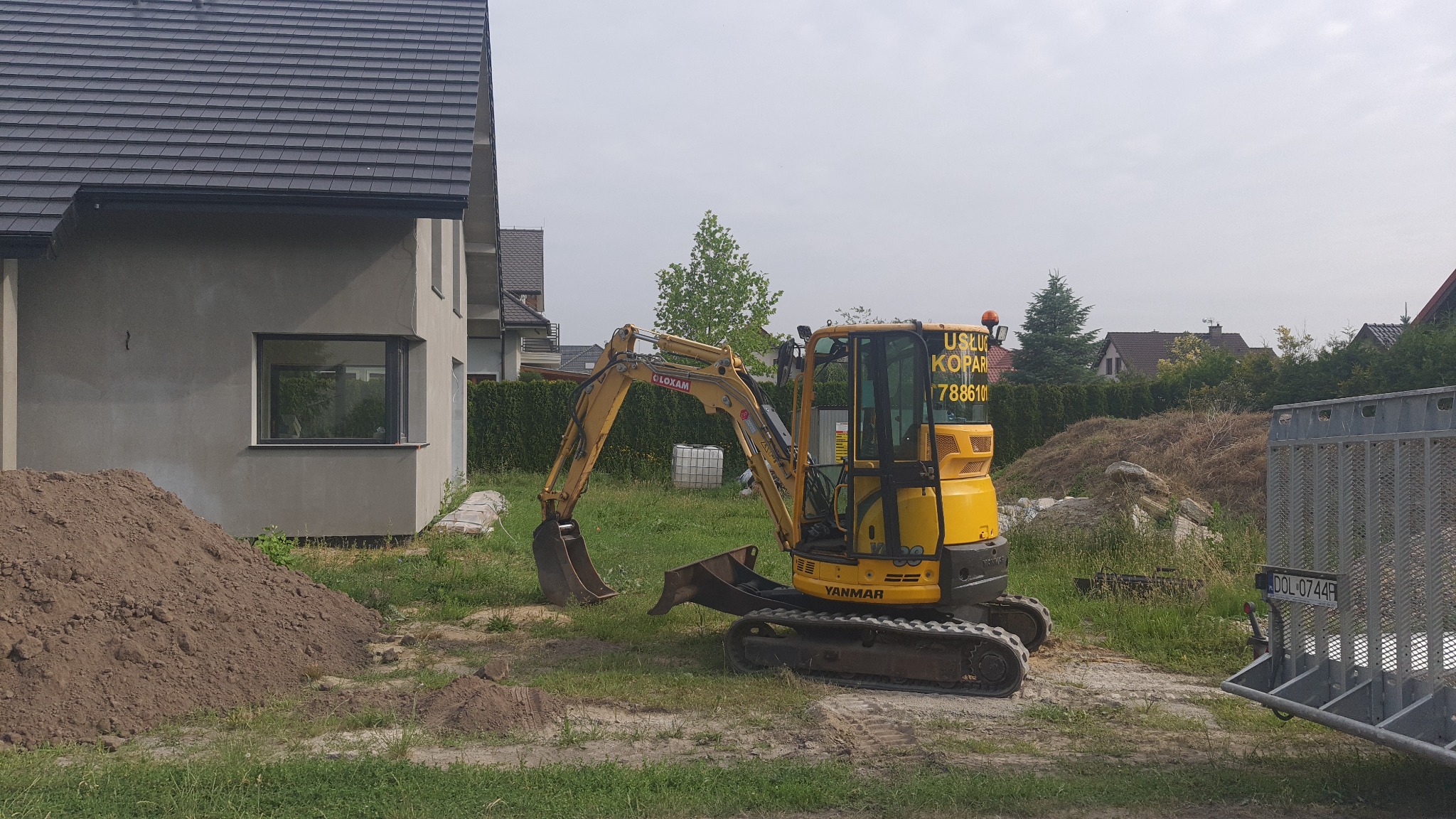 Żółta mini koparka Yanmar na trawiastym terenie budowy, obok sterta ziemi i budynek w surowym stanie, z widoczną przyczepką z tyłu i numerem telefonu na kabinie.