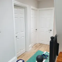 Korytarz po remoncie z trzema białymi drzwiami z czarnymi klamkami, widoczna podłoga z paneli, czarna balustrada schodów, mop i para butów na zielonej macie ochronnej.