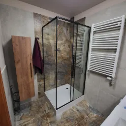 Nowoczesna łazienka z kabiną prysznicową typu walk-in z czarną ramą, drewnianą szafką wiszącą, grzejnikiem drabinkowym i płytkami imitującymi kamień w odcieniach brązu i szarości.