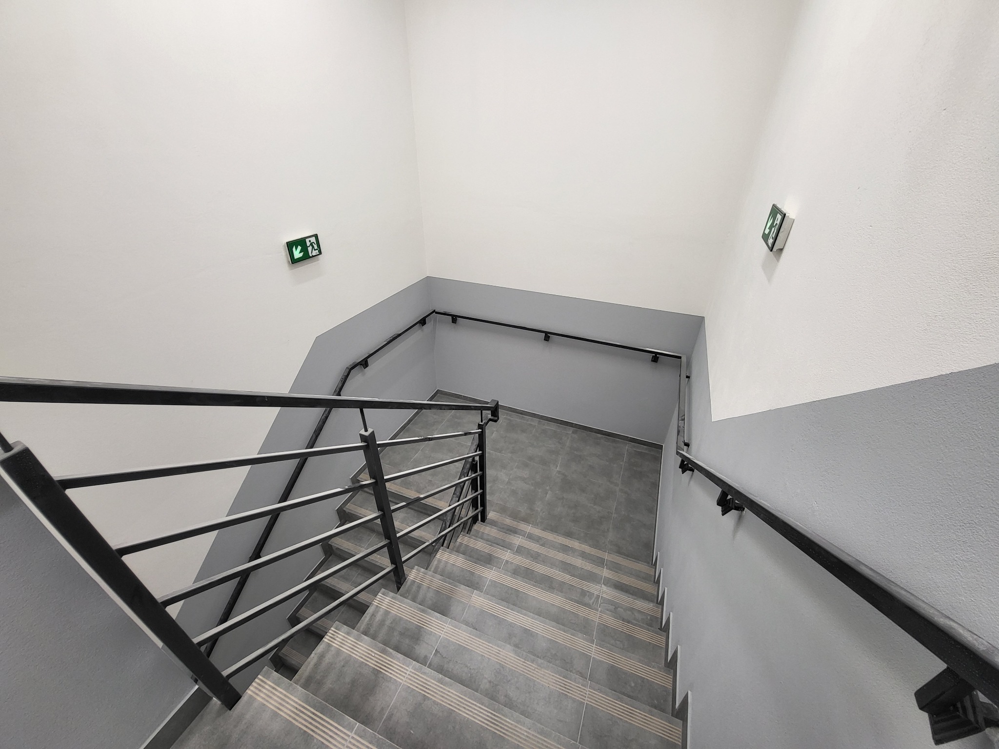Klatka schodowa z szarymi płytkami antypoślizgowymi na schodach, pomalowana na biało i szaro, z czarną metalową balustradą i oznaczeniami drogi ewakuacyjnej na ścianach.