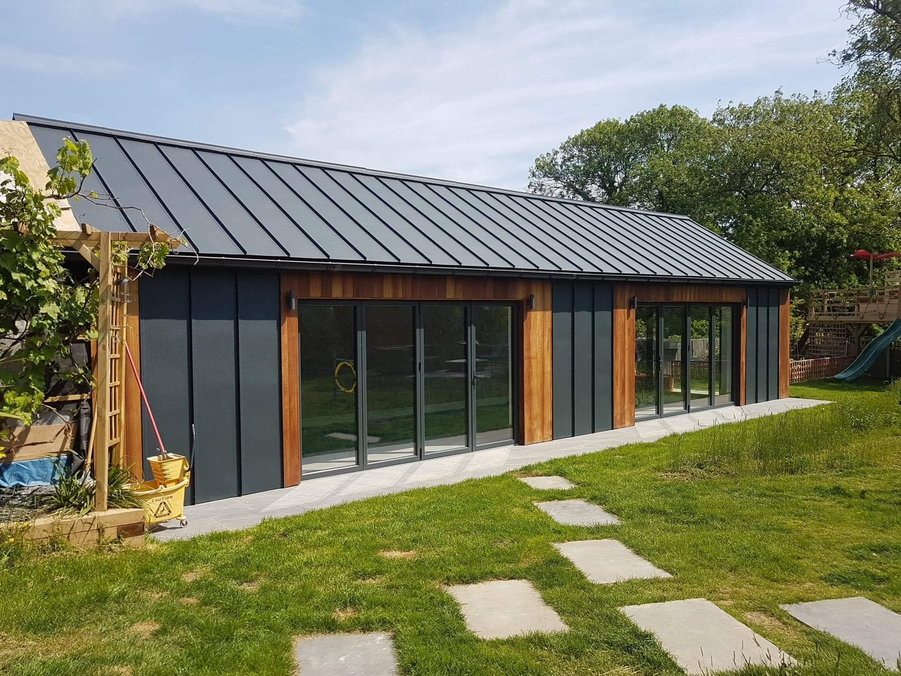 Nowoczesny budynek gospodarczy z szarą, metalową blachą na dachu, drewnianą elewacją i dużymi, przeszklonymi drzwiami przesuwnymi, widok z zewnątrz, trawnik z kamiennymi płytami ścieżki.