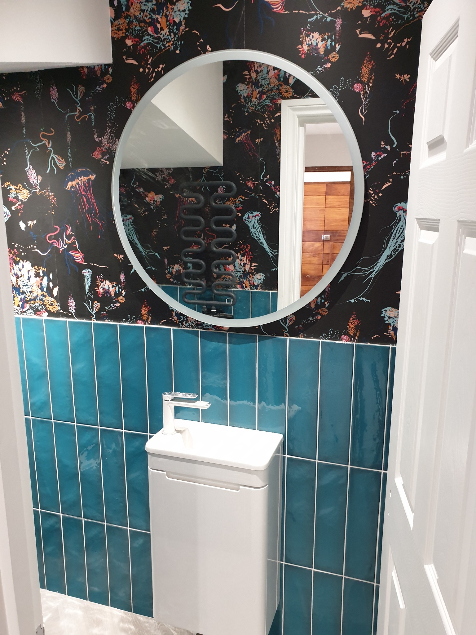 Nowoczesna łazienka z umywalką na szafce, turkusowymi płytkami wertykalnymi i tapetą w meduzy, okrągłe lustro odbija drewniane panele na ścianie w sąsiednim pomieszczeniu.