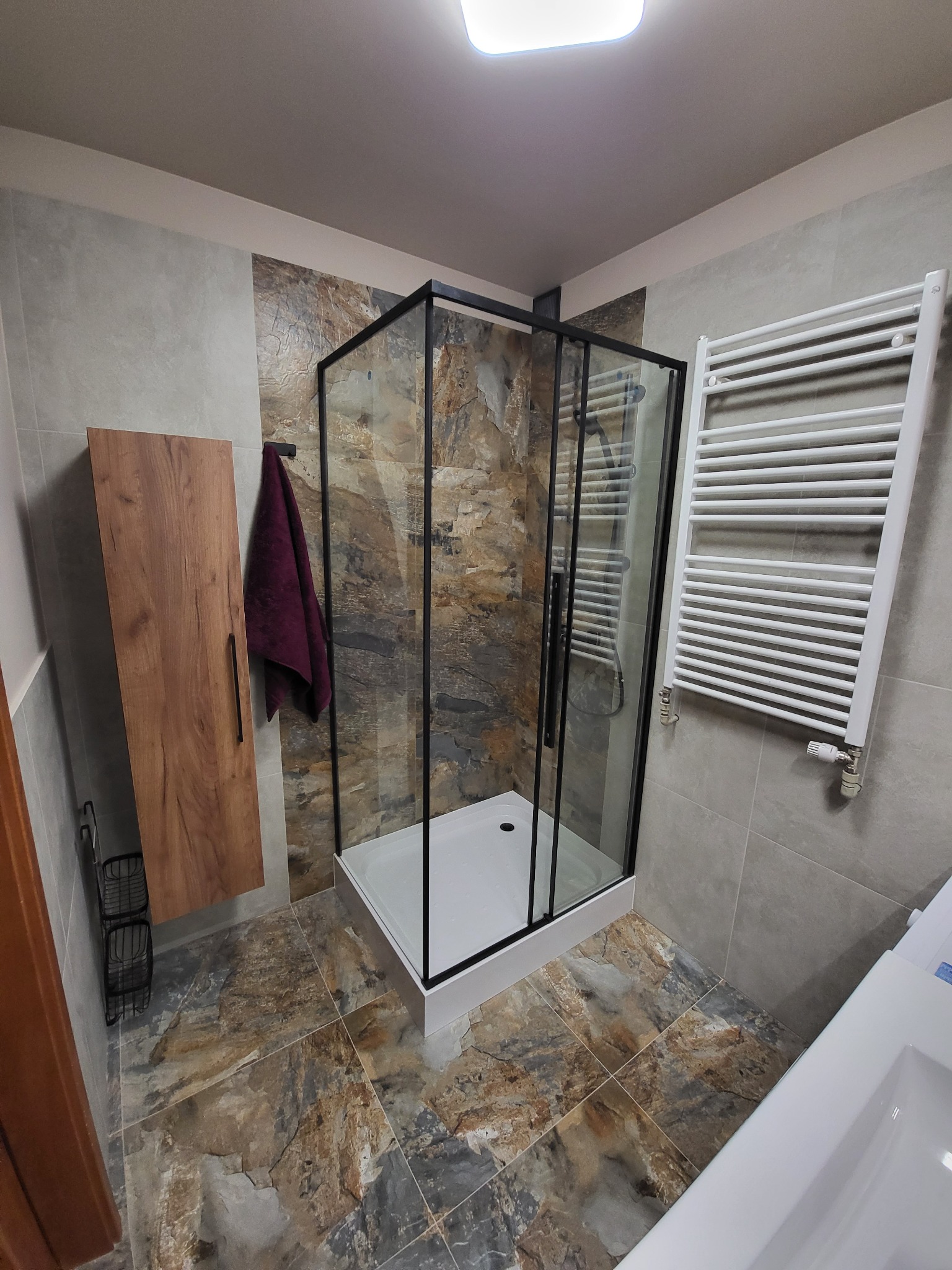 Nowoczesna łazienka z kabiną prysznicową typu walk-in z czarną ramą, drewnianą szafką wiszącą, grzejnikiem drabinkowym i płytkami imitującymi kamień w odcieniach brązu i szarości.