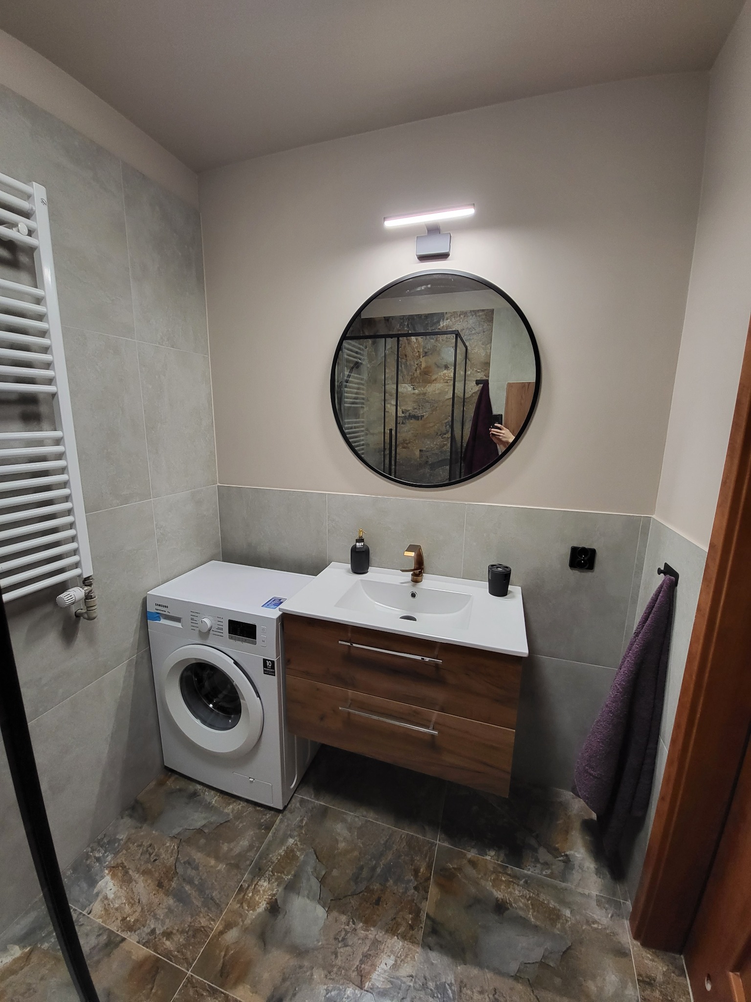 Nowoczesna łazienka z szarymi płytkami, okrągłym lustrem, drewnianą szafką pod umywalką z baterią w kolorze złota oraz pralką Samsung obok, widok na kabinę prysznicową w odbiciu lustra.