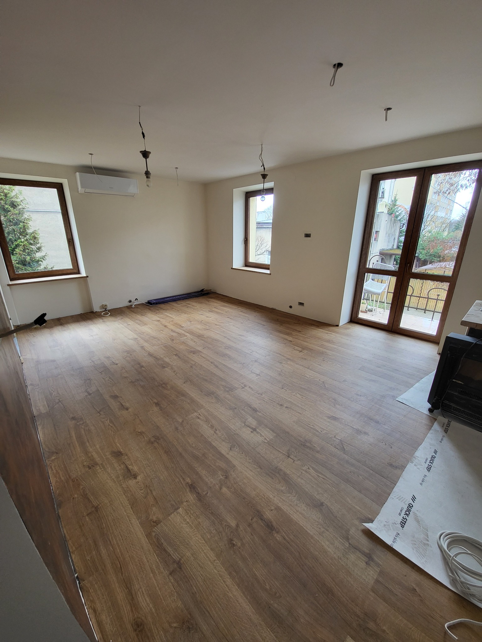 Wnętrze pokoju po ułożeniu paneli podłogowych w odcieniach brązu, widoczne okna z drewnianą ramą, sufit z wystającymi kablami od oświetlenia oraz klimatyzator, na podłodze leży folia pod panele.