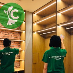 CLEANHOUSE - Dwie osoby w zielonych koszulkach z napisem 'CLEANHOUSE TEAM' czyszczą półki w garderobie z oświetleniem LED, widoczny fragment ściany z cegły.
