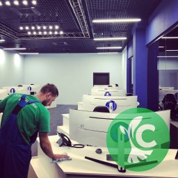 CLEANHOUSE - Mężczyzna w zielonej koszulce i niebieskich ogrodniczkach przeciera wilgotną szmatką białe biurko w nowoczesnym biurze z rzędami monitorów i metalową siatką pod sufitem.