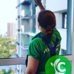 CLEANHOUSE - Kobieta w zielonej koszulce i niebieskich spodniach roboczych czyści okno w nowoczesnym budynku, widok zza pleców na miasto w tle.