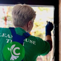 CLEANHOUSE - Osoba w niebieskich rękawicach maluje ramę okienną na czarno, widok od tyłu, logo firmy na koszulce.