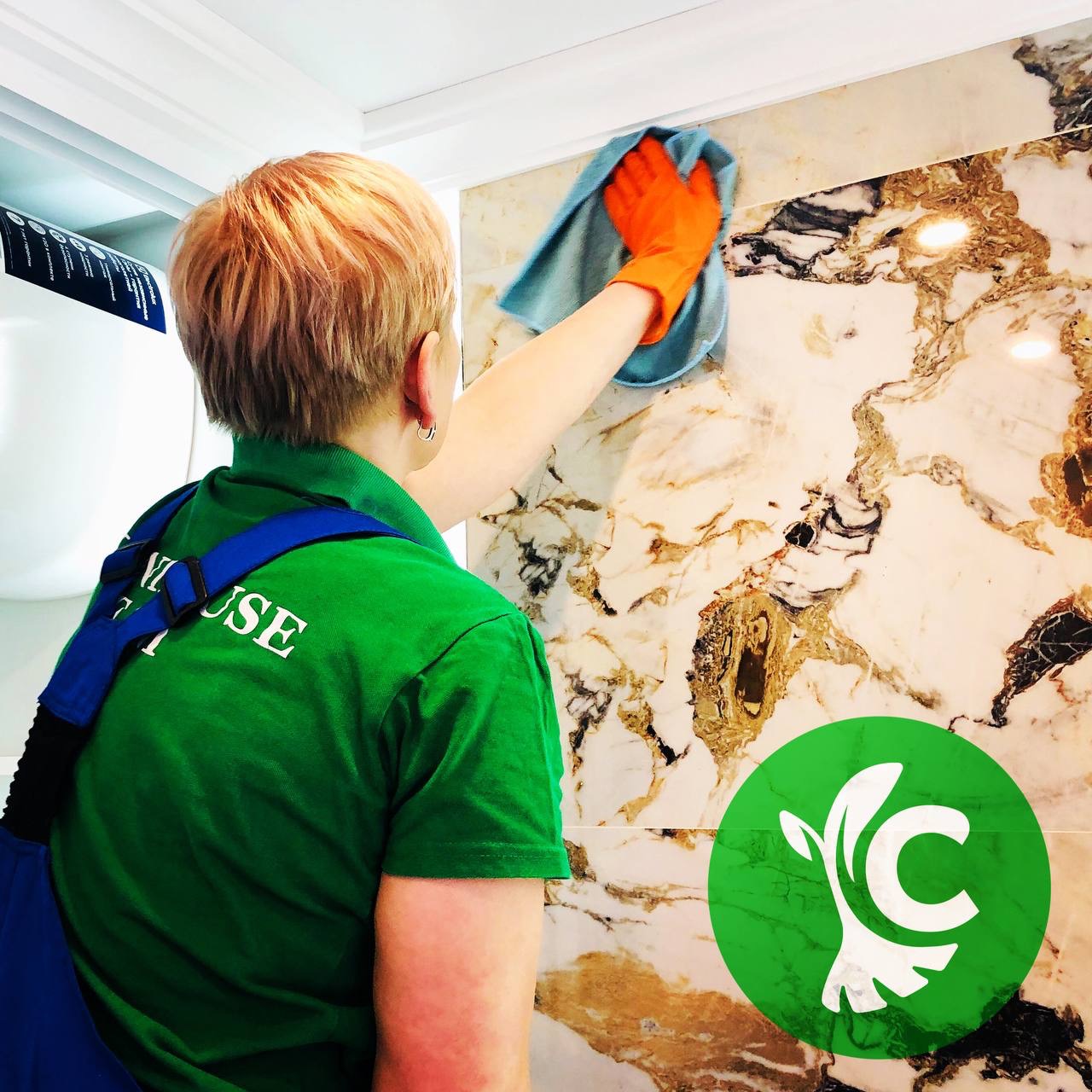 Osoba w zielonej koszulce i pomarańczowych rękawicach czyści marmurową ścianę niebieską ściereczką; widoczny fragment logotypu firmy nałożony na zdjęcie.