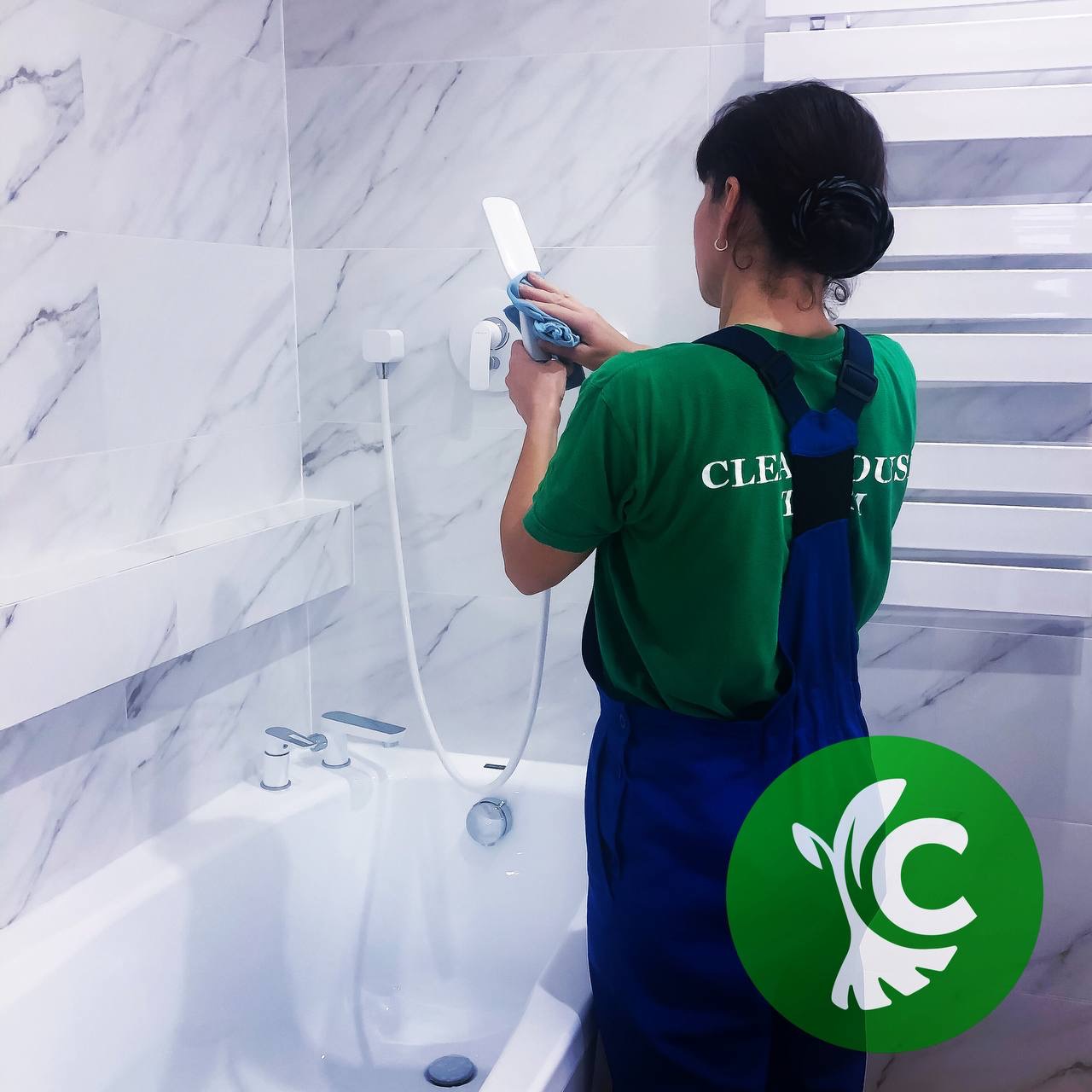 Pracownica w zielonej koszulce i granatowym kombinezonie czyści słuchawkę prysznicową w łazience wyłożonej białym marmurem, używając niebieskiej ściereczki.