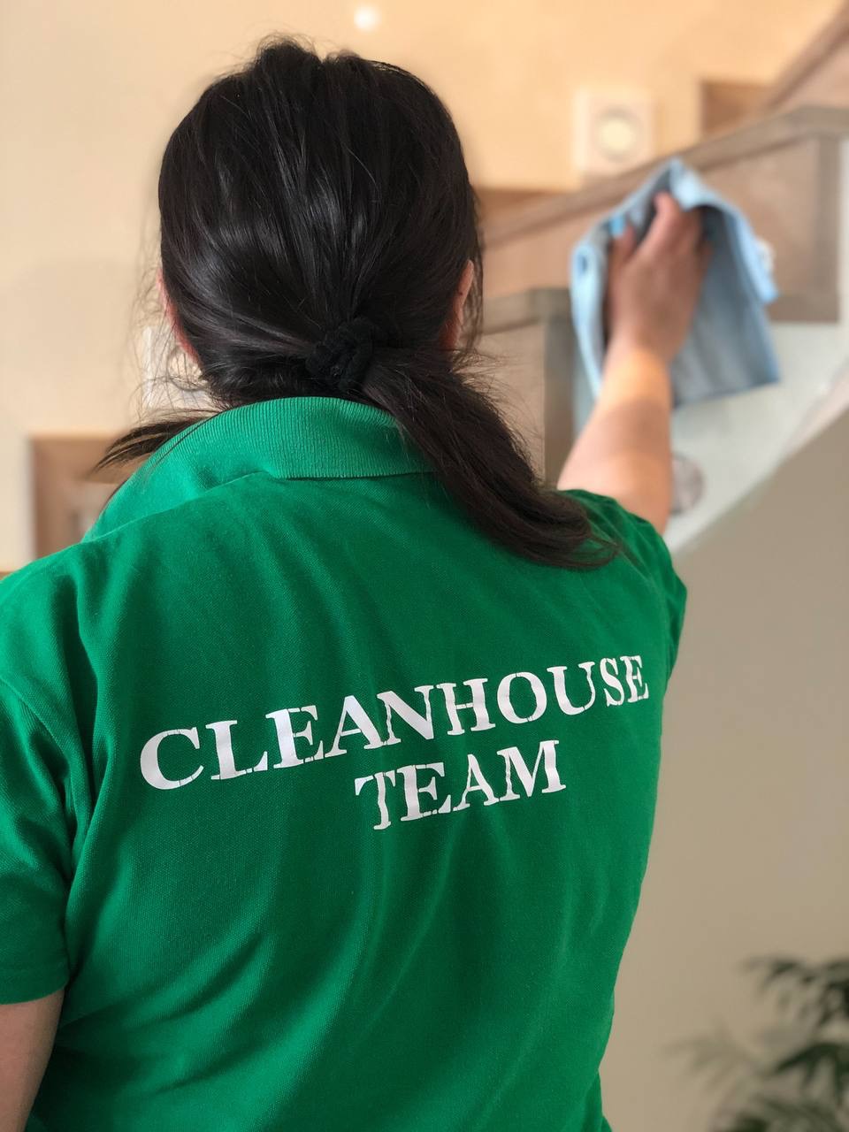Pracownica w zielonej koszulce z napisem 'CLEANHOUSE TEAM' czyści jasne, drewniane schody za pomocą niebieskiej ściereczki.