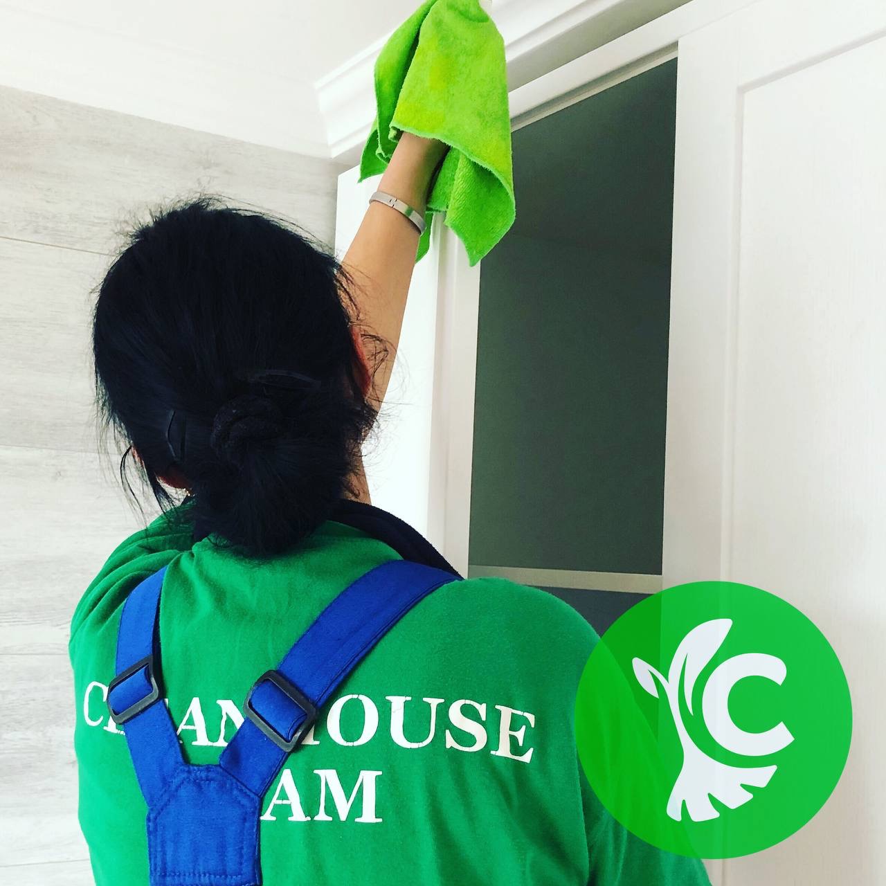 Pracownica w zielonej koszulce i niebieskich szelkach przeciera wilgotną, zieloną ściereczką górną krawędź białych drzwi w pomieszczeniu.