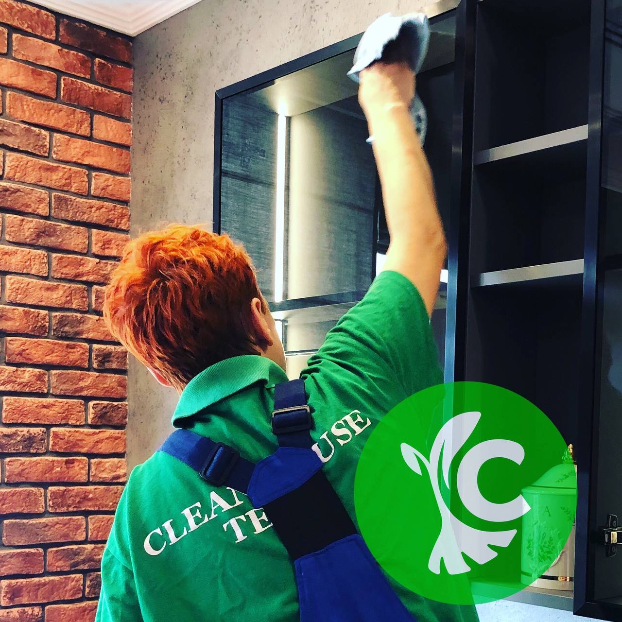 Pracownik w zielonej koszulce z napisem 'CLEAN TE USE' czyści szmatką półki w ciemnej szafce, widoczna ceglana ściana.