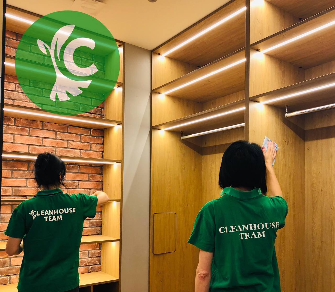 Dwie osoby w zielonych koszulkach z napisem 'CLEANHOUSE TEAM' czyszczą półki w garderobie z oświetleniem LED, widoczny fragment ściany z cegły.