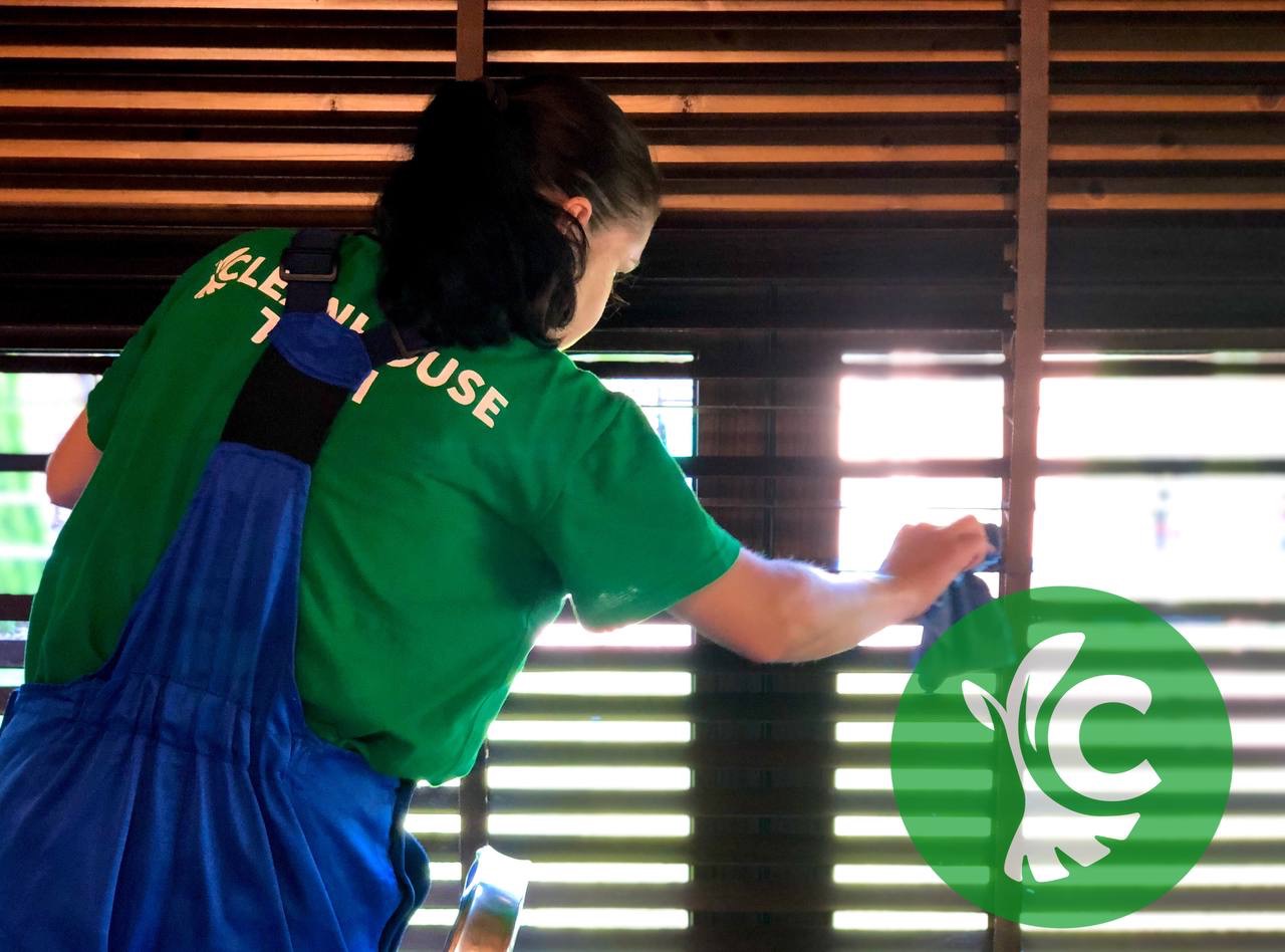 Pracownica w zielonej koszulce i niebieskich ogrodniczkach czyści żaluzje w pomieszczeniu w Warszawie, używając niebieskiej ściereczki.