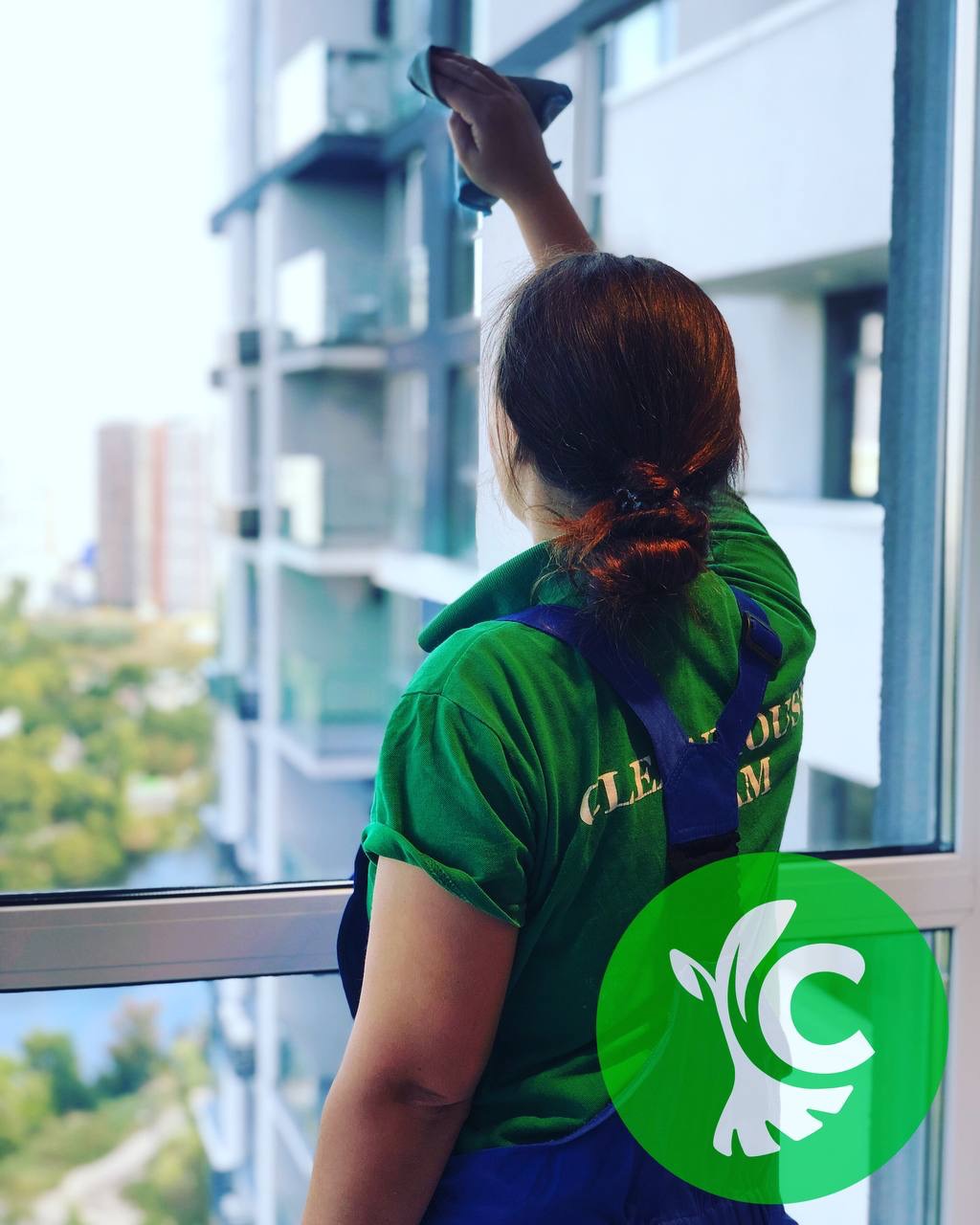 Kobieta w zielonej koszulce i niebieskich spodniach roboczych czyści okno w nowoczesnym budynku, widok zza pleców na miasto w tle.