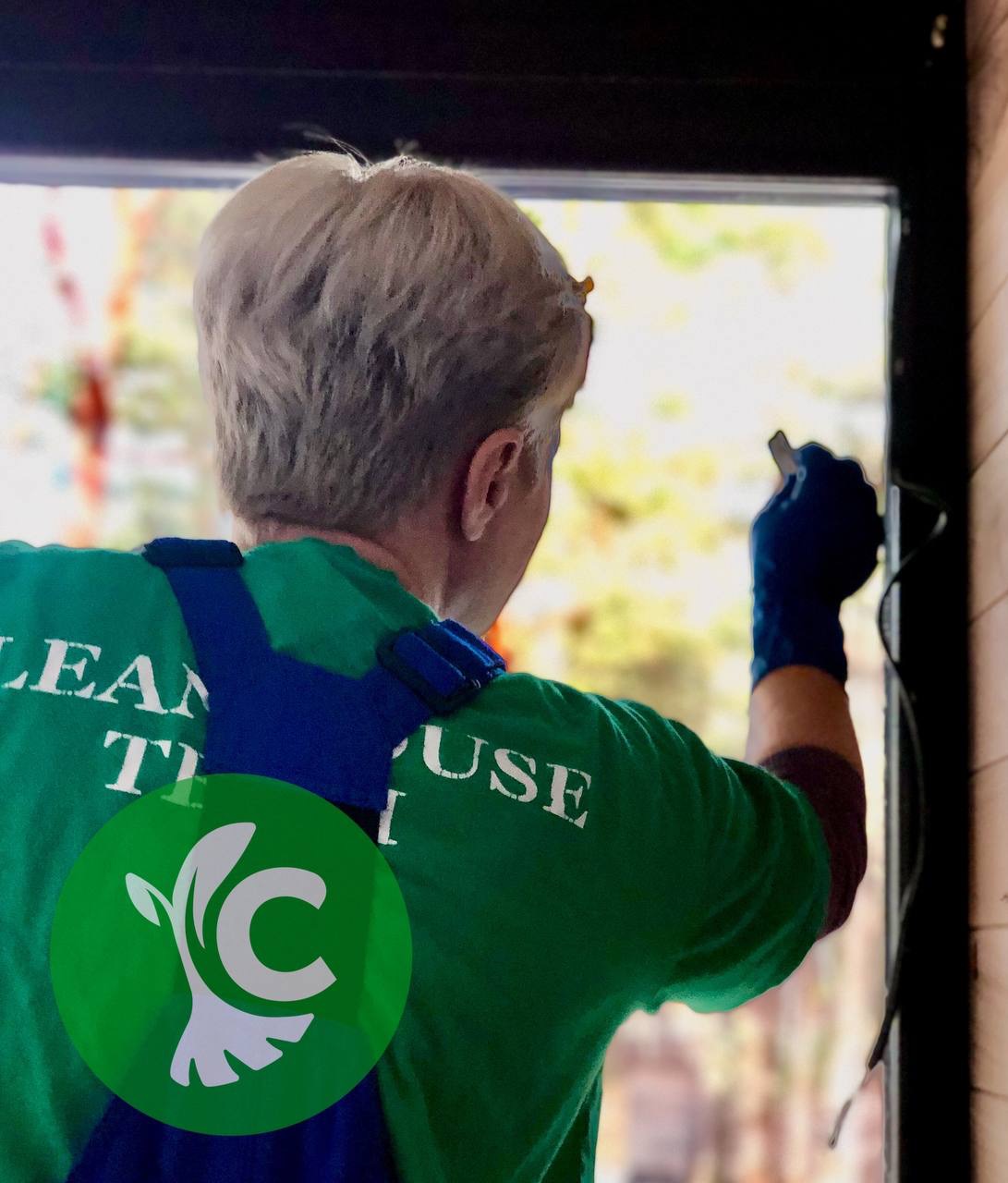 Osoba w niebieskich rękawicach maluje ramę okienną na czarno, widok od tyłu, logo firmy na koszulce.